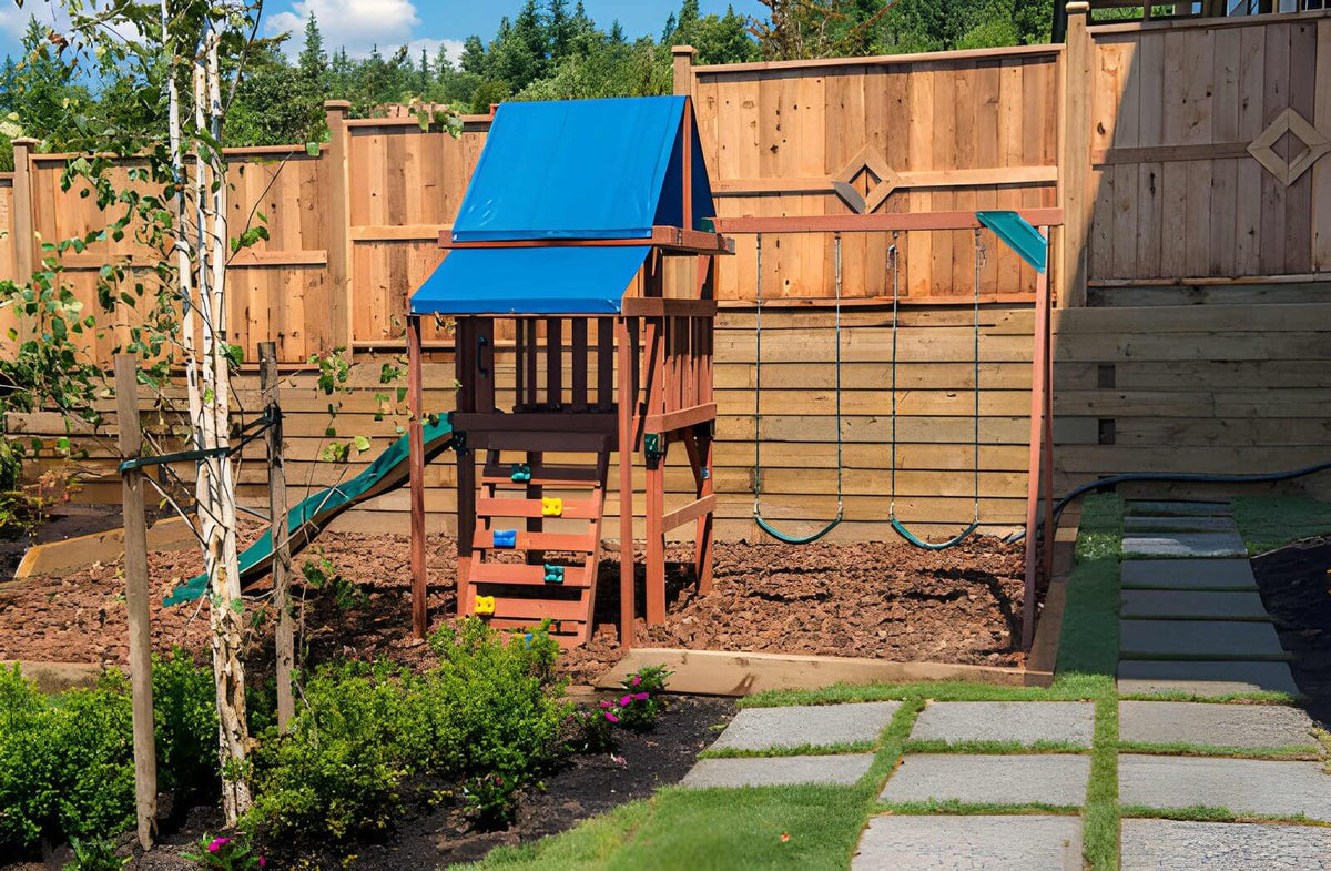 Simple backyard playground with wooden playset, slide and swings on mulch, surrounded by fence and plants.